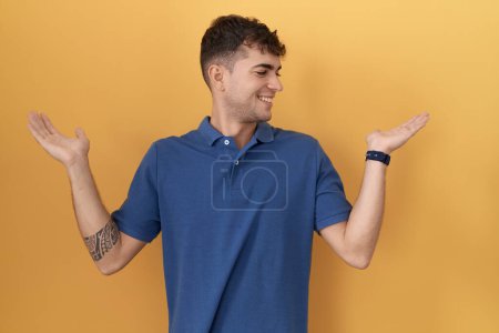 Photo for Young hispanic man standing over yellow background smiling showing both hands open palms, presenting and advertising comparison and balance - Royalty Free Image