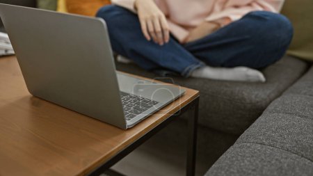 Téléchargez les photos : Une jeune femme s'assoit tranquillement sur un canapé gris près d'un ordinateur portable ouvert dans un cadre confortable de salon. - en image libre de droit