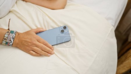 Foto de Primer plano de la mano de una mujer madura con brazalete sosteniendo un smartphone en una cama en una habitación bien iluminada, lo que sugiere un ambiente interior relajado. - Imagen libre de derechos