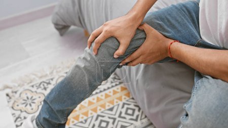 Téléchargez les photos : Jeune homme tenant le genou dans la douleur assis sur un tapis à motifs géométriques à la maison, transmettant un besoin de soins médicaux ou de récupération des blessures. - en image libre de droit