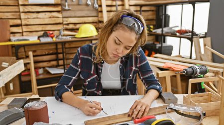 Attractive young hispanic woman carpenter, dressed for safety with glasses, skillfully jots down notes in her carpentry workshop