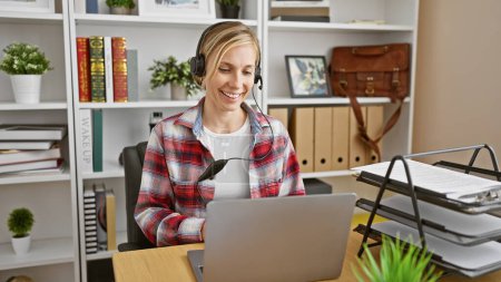 Photo for A smiling caucasian woman with a headset works at her laptop in a modern home office. - Royalty Free Image