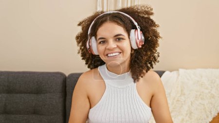 Téléchargez les photos : Jeune femme hispanique aux cheveux bouclés portant des écouteurs, souriant à l'intérieur à la maison sur un canapé. - en image libre de droit