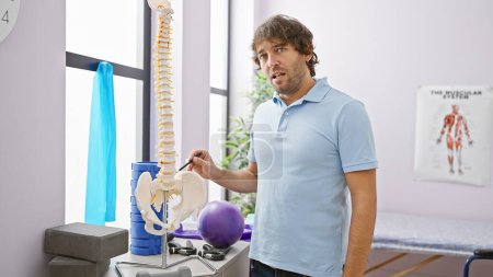 Téléchargez les photos : Bel homme barbu pointant vers le modèle de colonne vertébrale dans une salle de clinique de réadaptation avec équipement d'exercice. - en image libre de droit