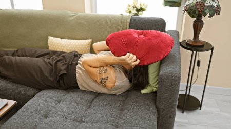 Téléchargez les photos : Femme hispanique reposant sur le canapé face cachée avec coussin rouge dans le salon confortable - en image libre de droit