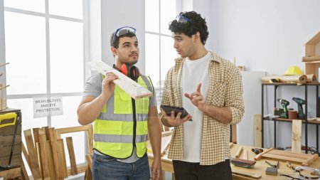 Photo for Two men engaged in discussion over a tablet in a bright carpentry workshop filled with tools - Royalty Free Image