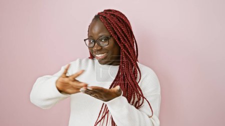 Una mujer negra alegre con trenzas hace un gesto de dinero juguetón contra una pared rosa