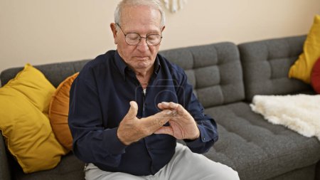 Weathered man with white hair stretching achy hand, looking serious while relaxing on room's sofa, suffering from arthritis at home