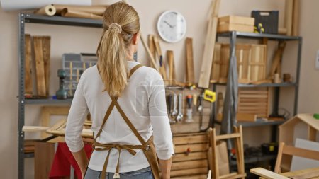 Téléchargez les photos : Vue arrière d'une femme blonde debout dans un atelier de menuiserie bien organisé, entouré de matériaux et d'outils en bois. - en image libre de droit