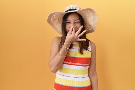 Téléchargez les photos : Femme chinoise d'âge moyen portant un chapeau d'été sur fond jaune sentant quelque chose de puant et dégoûtant, odeur intolérable, retenant la respiration avec les doigts sur le nez. mauvaise odeur - en image libre de droit