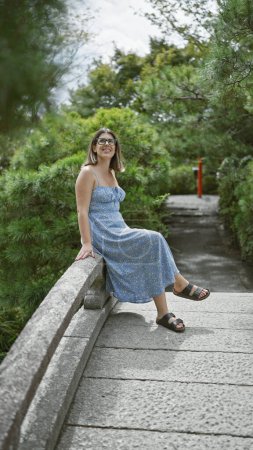 Téléchargez les photos : Belle jeune femme hispanique portant des lunettes souriant assis sur un pont au parc japonais - en image libre de droit