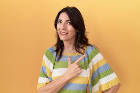 Photo for Middle age hispanic woman standing over yellow background cheerful with a smile of face pointing with hand and finger up to the side with happy and natural expression on face - Royalty Free Image