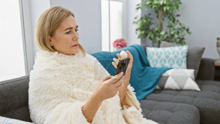 Foto de Una mujer rubia de mediana edad examina la medicación mientras está sentada en un sofá en una sala de estar moderna. - Imagen libre de derechos