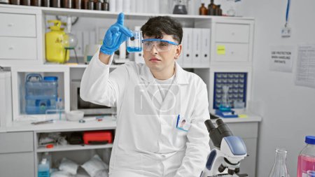 Foto de Un joven con una bata de laboratorio examina un líquido azul en un ambiente de laboratorio, rodeado de equipo científico. - Imagen libre de derechos