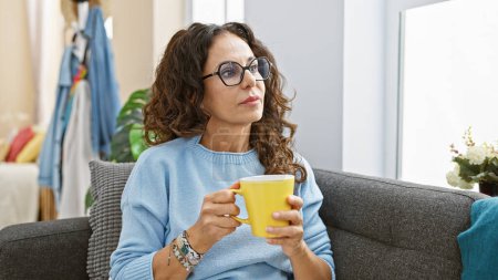 Téléchargez les photos : Femme hispanique mature dans des lunettes s'assoit soigneusement sur un canapé à la maison, tenant une tasse jaune. - en image libre de droit