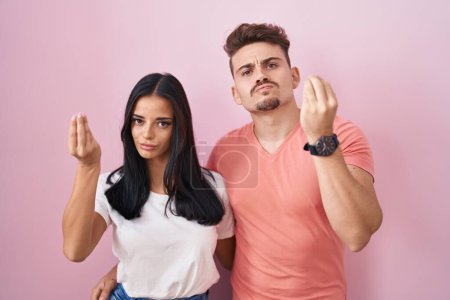 Foto de Young hispanic couple standing over pink background doing italian gesture with hand and fingers confident expression - Imagen libre de derechos