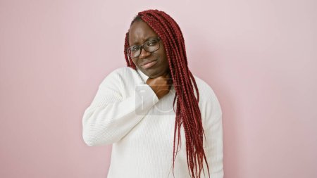 Foto de Mujer afroamericana con trenzas con gafas y un suéter blanco sobre un fondo rosa se ve incómoda sosteniendo su garganta. - Imagen libre de derechos