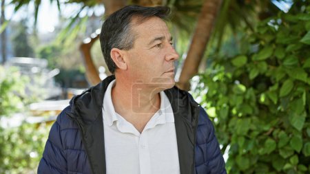 Photo for Contemplative middle-aged man outdoors, in casual attire, standing surrounded by greenery in a park. - Royalty Free Image