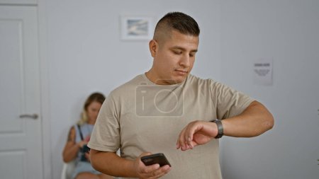 Téléchargez les photos : Beau jeune homme latino absorbé par les textos tech-savvy sur son smartphone, regardant avec ferveur sa montre en attendant patiemment dans une salle intérieure animée, immergée dans une connexion numérique. - en image libre de droit