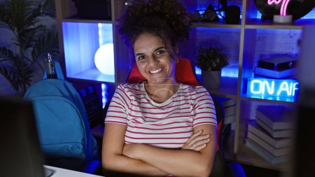 Smiling young hispanic woman with curly hair, arms crossed, in dark modern gaming room at night.