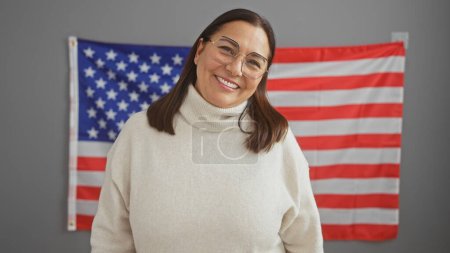 Smiling middle-aged hispanic woman in a white sweater standing before an american flag in an office setting