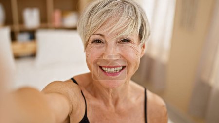 Smiling middle-aged hispanic woman takes a selfie in her cozy home bedroom, showcasing her short grey hair.