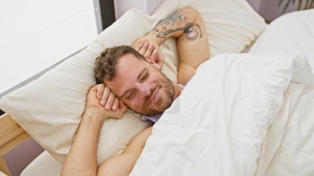 Téléchargez les photos : Un jeune homme souriant et tatoué s'allongeant confortablement au lit, incarnant détente et contentement dans un cadre de chambre confortable. - en image libre de droit