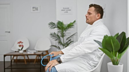 Photo for Hispanic man in white lab coat sitting thoughtfully in a modern clinic interior with a stethoscope and indoor plants. - Royalty Free Image