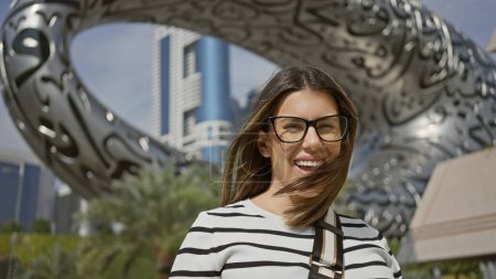 Téléchargez les photos : Une jeune femme souriante avec des lunettes se tient devant l'architecture futuriste dans dubai, incarnant un design moderne - en image libre de droit