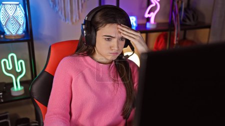 Foto de Una joven ansiosa con auriculares en una sala de juegos por la noche enfocada en la pantalla de su computadora. - Imagen libre de derechos