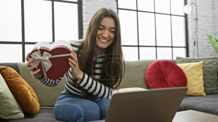 Photo for A cheerful young woman holding a heart-shaped gift box while sitting on a sofa in a well-lit living room, interacting with a laptop. - Royalty Free Image