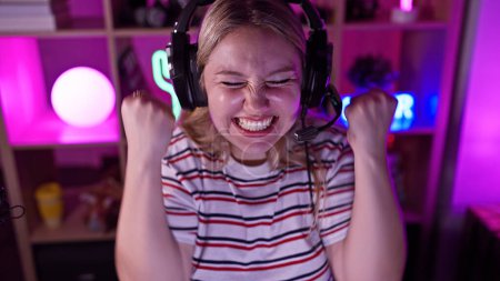 Excited young woman wearing headphones in a neon-lit gaming room expressing joy