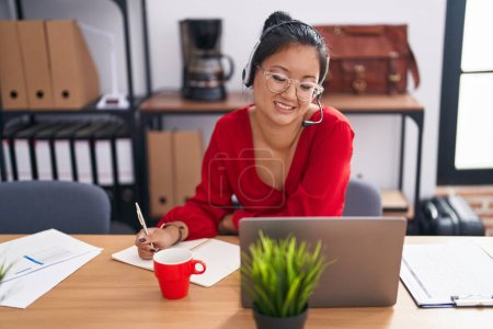 Photo for Young chinese woman call center agent writing on notebook working at office - Royalty Free Image