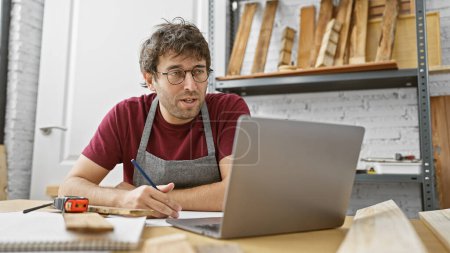 Photo for Bearded craftsman in apron working on laptop in a woodshop, surrounded by timber and tools. - Royalty Free Image