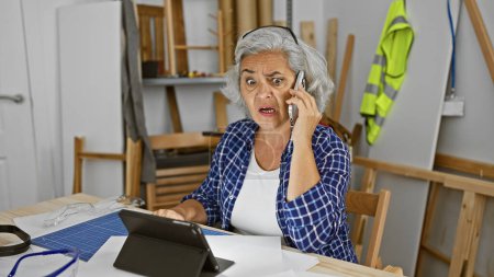 Photo for Surprised woman speaking on phone at carpentry workshop beside tablet and blueprints. - Royalty Free Image