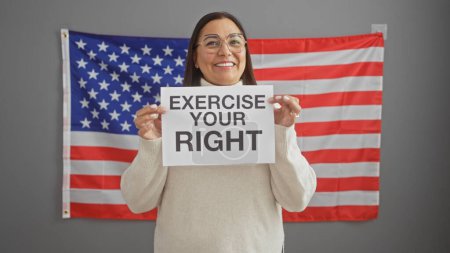 Foto de Mujer hispana sonriente sosteniendo un cartel 'ejerce tu derecho' frente a una bandera americana en el interior - Imagen libre de derechos