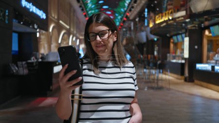 Eine junge erwachsene hispanische Frau benutzt ein Smartphone in einem modernen Luxus-Kreuzfahrtschiff und lächelt, während sie ihren Urlaub genießt.