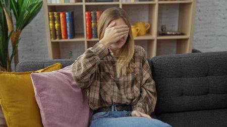 Gestresste junge Frau, die das Gesicht mit der Hand bedeckt, sitzt auf einer grauen Couch in einem gemütlichen Wohnzimmer zu Hause.
