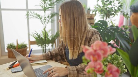 Téléchargez les photos : Une jeune femme blonde prend des notes dans un magasin de fleurs rempli de verdure, travaillant sur un ordinateur portable dans un cadre intérieur. - en image libre de droit