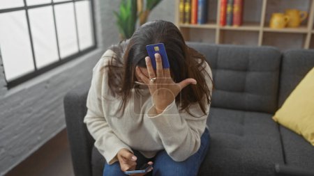 Foto de Mujer estresada sostiene tarjeta de crédito a la frente sentado en el sofá en un entorno interior de la casa con teléfono inteligente. - Imagen libre de derechos