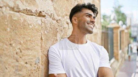 Photo for Handsome young hispanic man smiling outdoors in a sunny urban setting, wearing a casual white shirt and leaning on a stone wall. - Royalty Free Image