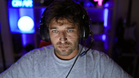 Handsome hispanic man with beard and green eyes wearing headphones in a dark gaming room at night