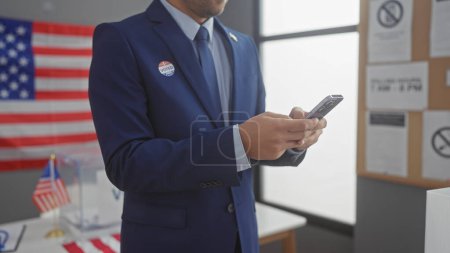 A young man with a beard, dressed in a suit, checks his phone at an american electoral center, with a flag and voting booths.