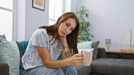 Photo for A thoughtful young woman sits on a sofa in a cozy living room, holding a mug, expressing contemplation. - Royalty Free Image