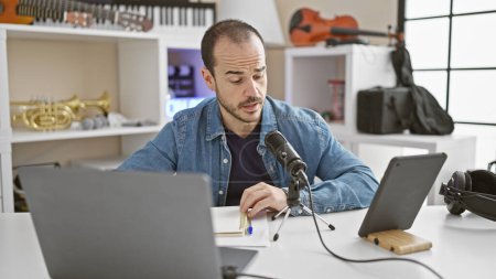 Hispanic bald man with beard recording in a modern music studio, using a microphone and digital tablet.