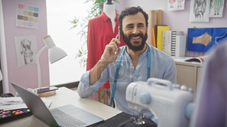 Téléchargez les photos : Un homme barbu souriant dans un magasin de tailleurs est au téléphone, entouré de matériel de couture, de tissu et de mannequin. - en image libre de droit