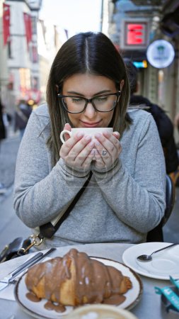 Téléchargez les photos : Une jeune femme dégustant du café et des croissants à une table de café extérieure dans une rue animée de la ville. - en image libre de droit