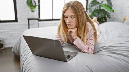 Téléchargez les photos : Jeune femme caucasienne avec des cheveux blonds utilisant intensément un ordinateur portable tout en étant couché sur un lit dans une chambre moderne lumineuse - en image libre de droit
