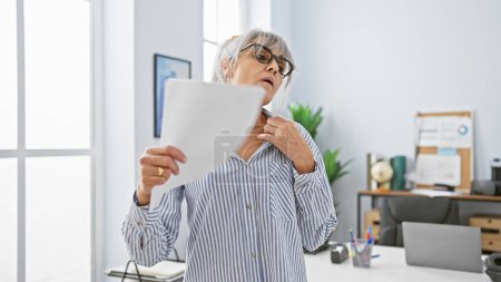 Femme hispanique mature avec des cheveux gris portant des lunettes dans un cadre de bureau regarde les documents