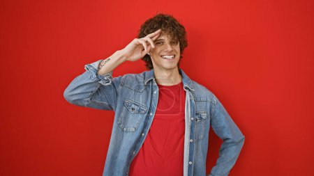 Foto de Un alegre joven hispano con barba y pelo rizado saluda juguetonamente sobre un vibrante fondo rojo, retratando la confianza casual. - Imagen libre de derechos